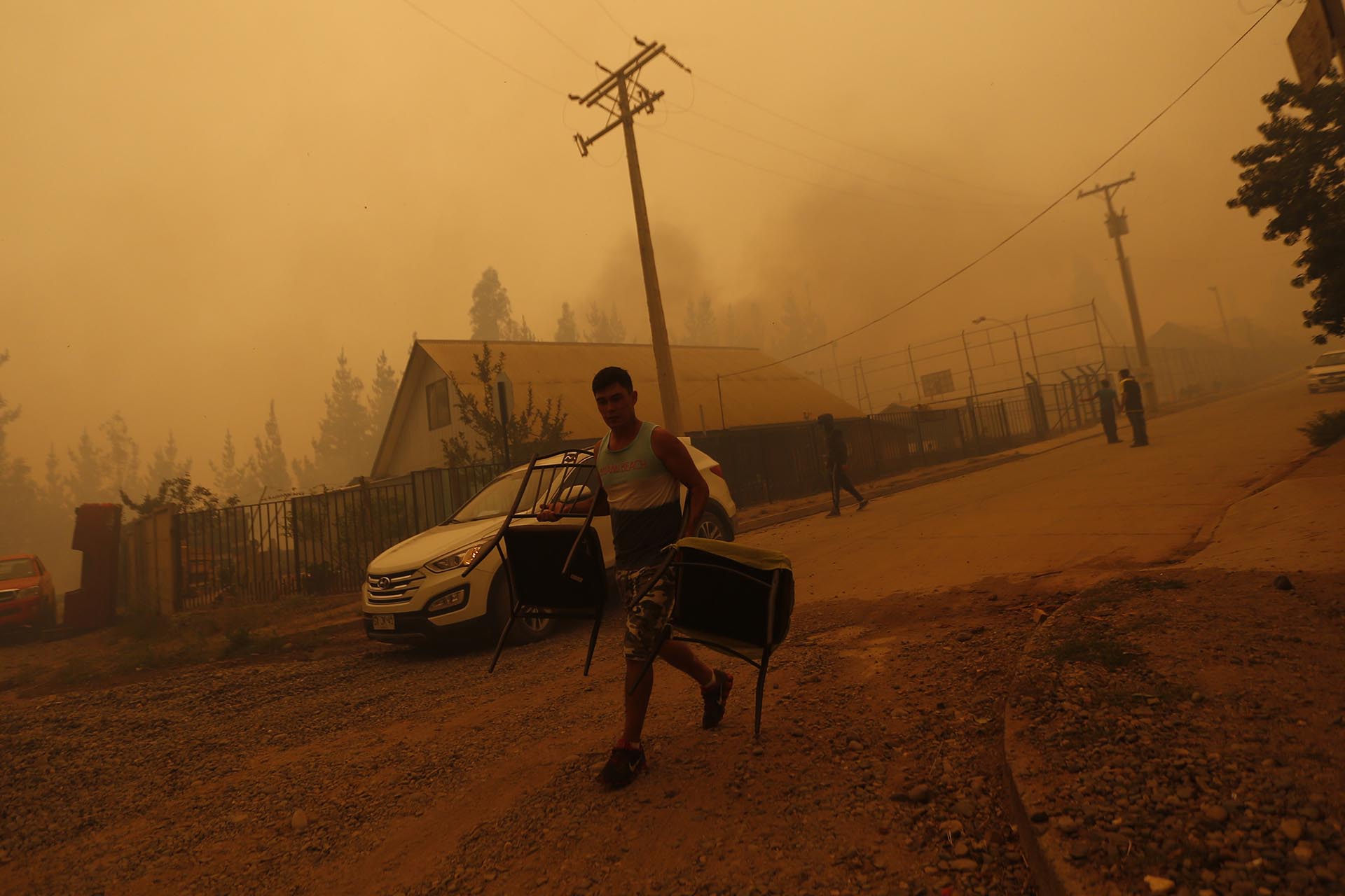 A young man evacuates carrying chairs in San Ramon after a forest fire devastated the nearby town of Santa Olga, 240 kilometres south of Santiago, on January 26, 2017. Six people -- among them four firefighters and two police -- have now been killed battling vast forest fires in central Chile, officials said Wednesday. Multiple blazes have ravaged 238,000 hectares (588,000 acres) and are growing, the National Forestry Corporation said in a statement. / AFP PHOTO / PABLO VERA LISPERGUER